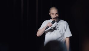 A white man with a large mustache performs stand-up comedy under a spotlight.